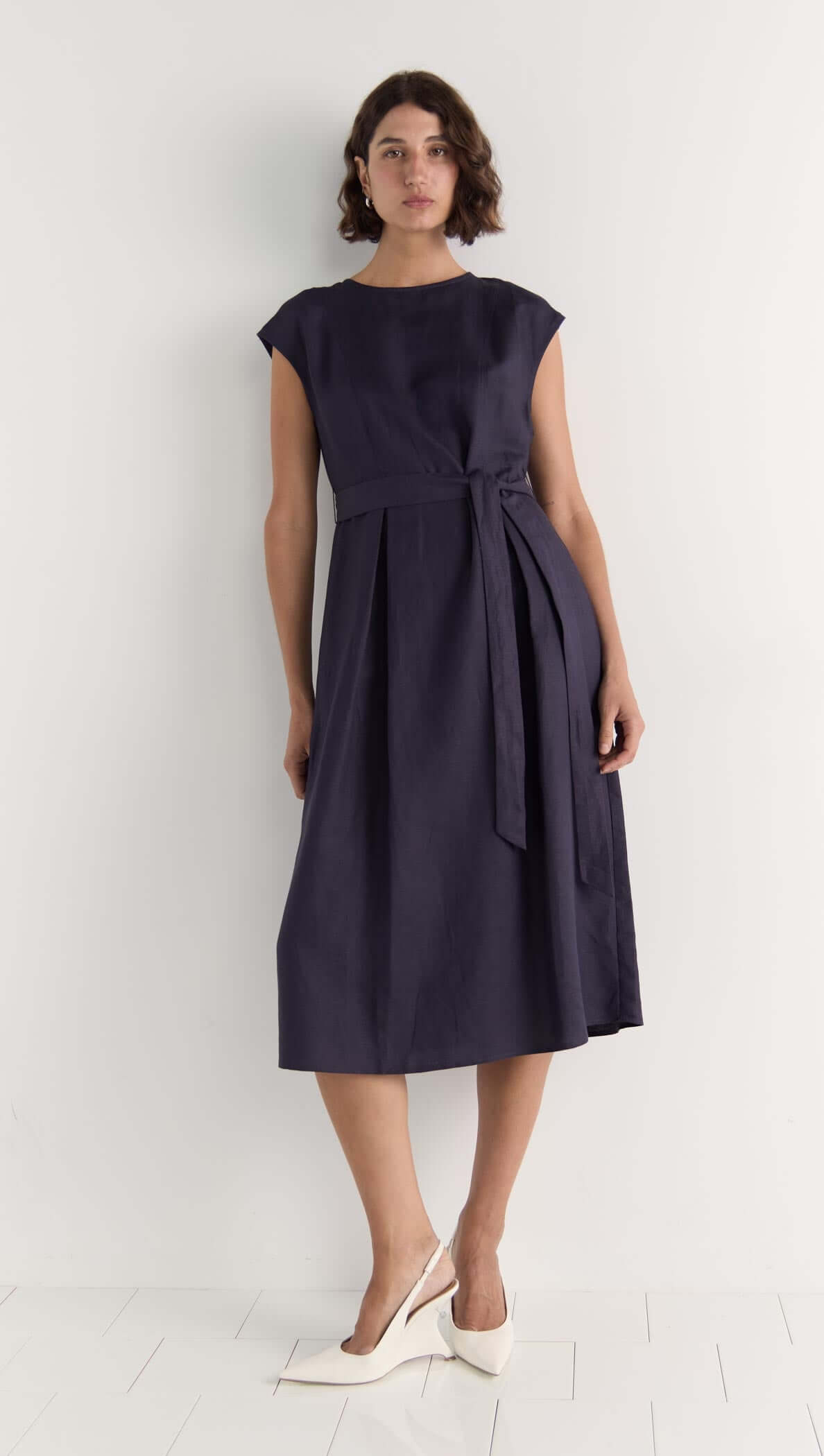 Model wearing the Rhea Midi Dress, a sleeveless deep navy dress with a self-tie belt and midi length.