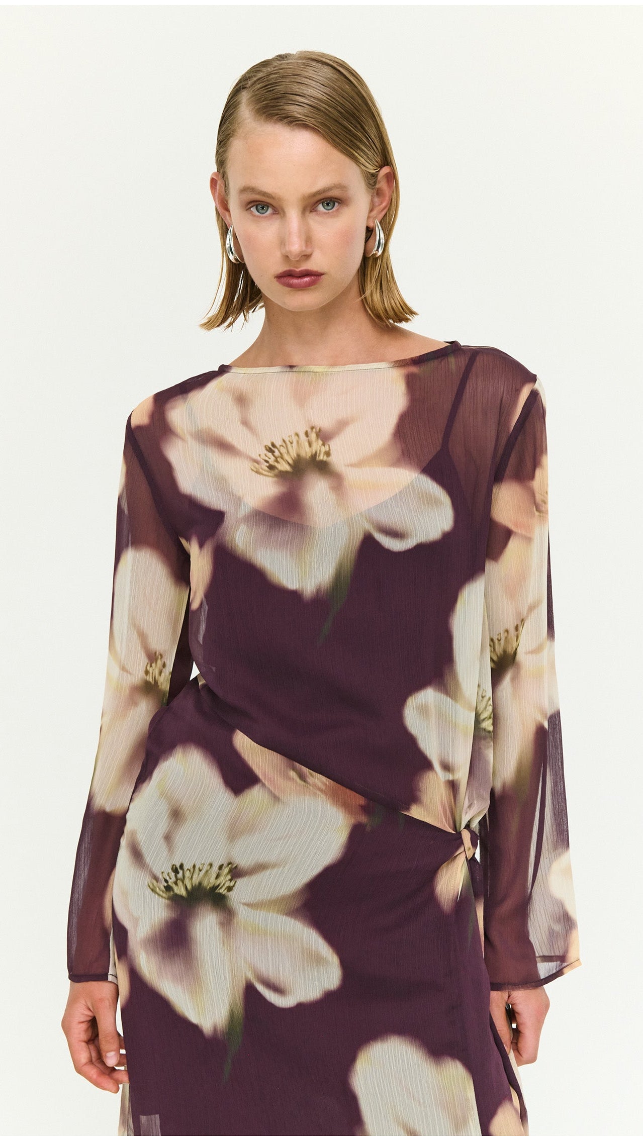 Model wearing a sheer floral maxi dress with tie detail, featuring large white flowers on a deep purple background.