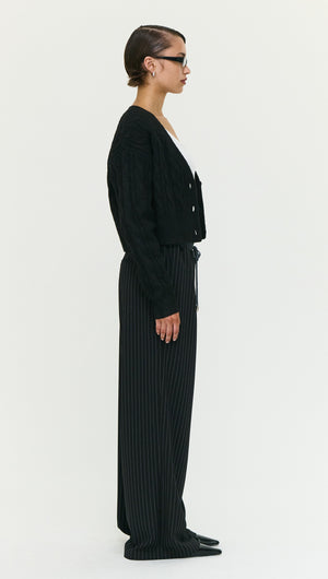 Model wearing a Black Cable Knit Cropped Cardigan paired with striped pants, showcasing a side view of the outfit.