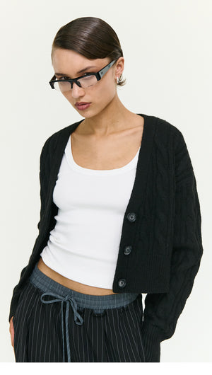 Stylish woman wearing a Black Cable Knit Cropped Cardigan over a white tank top, showcasing a modern fashion look.