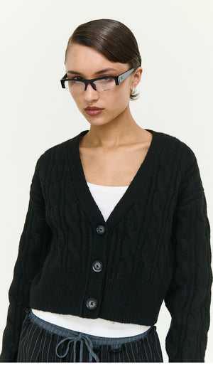 Model wearing a Black Cable Knit Cropped Cardigan with a V-neck and button closure, styled with glasses.