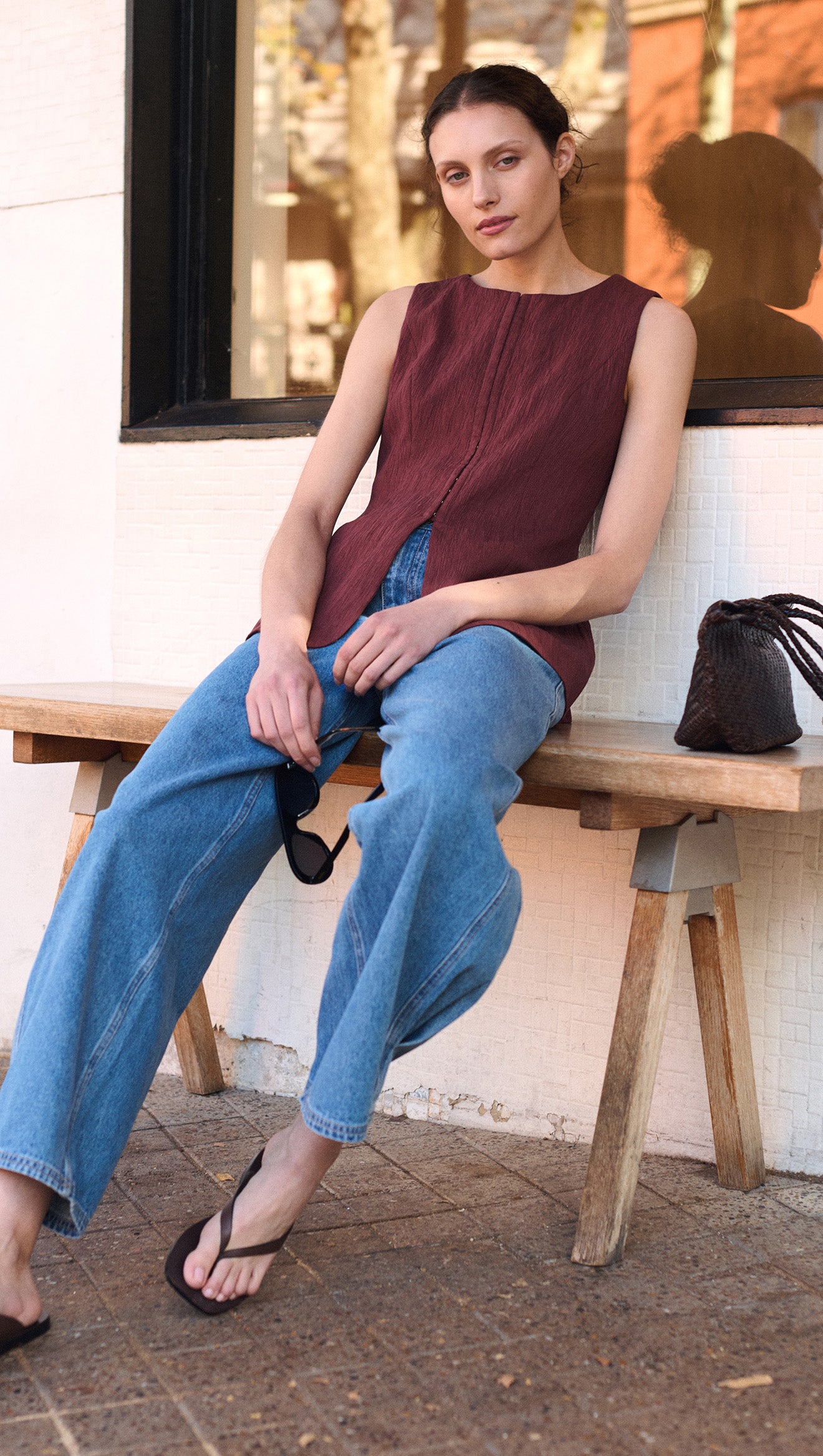 Model wearing Burgundy Round Neck Textured Vest with blue jeans, sitting on a bench outdoors.