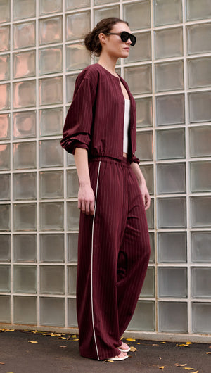 Model wearing Burgundy Pinstripe Relaxed Pant paired with a stylish jacket, showcasing modern fashion against a glass wall.
