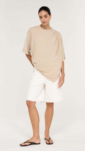 Asymmetric Draped Oversize Tee in Beige worn with white shorts and sandals, showcasing a relaxed fit and contemporary style.