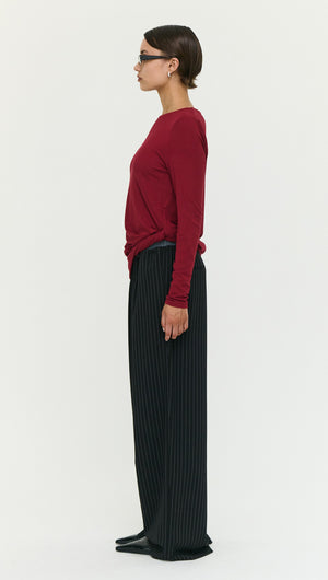 Model wearing Long Sleeve Jersey Twist Top in Merlot paired with black striped pants, showcasing stylish side view.