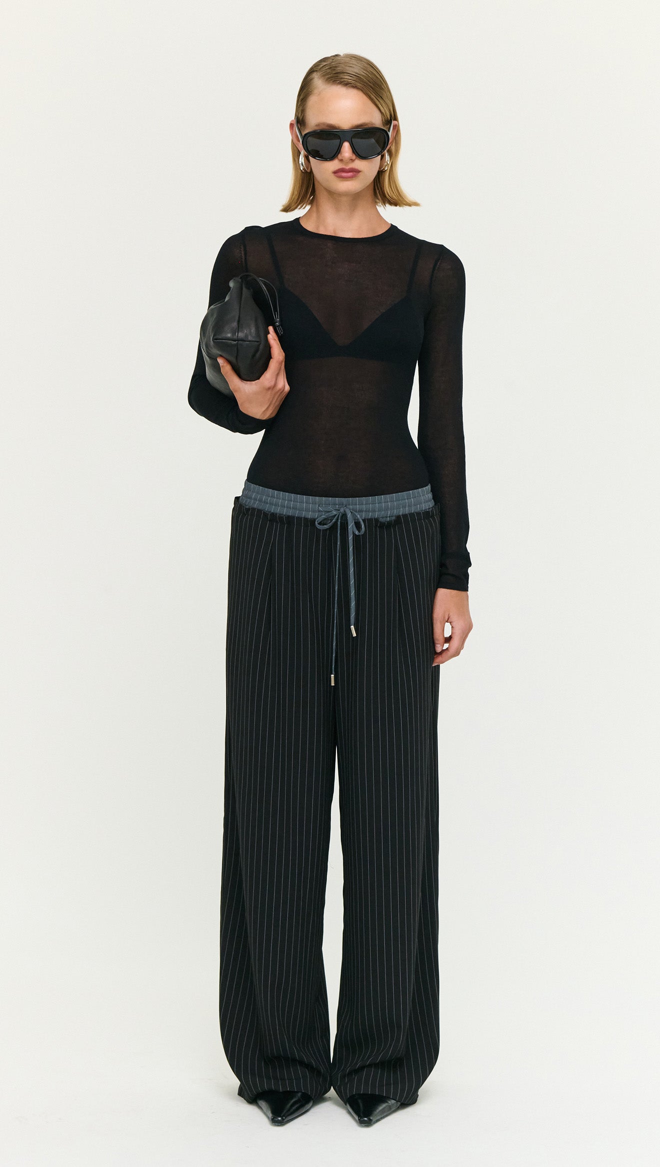 Model wearing Pinstripe Relaxed Drawstring Pants paired with a sheer black top and sunglasses, holding a small black bag.