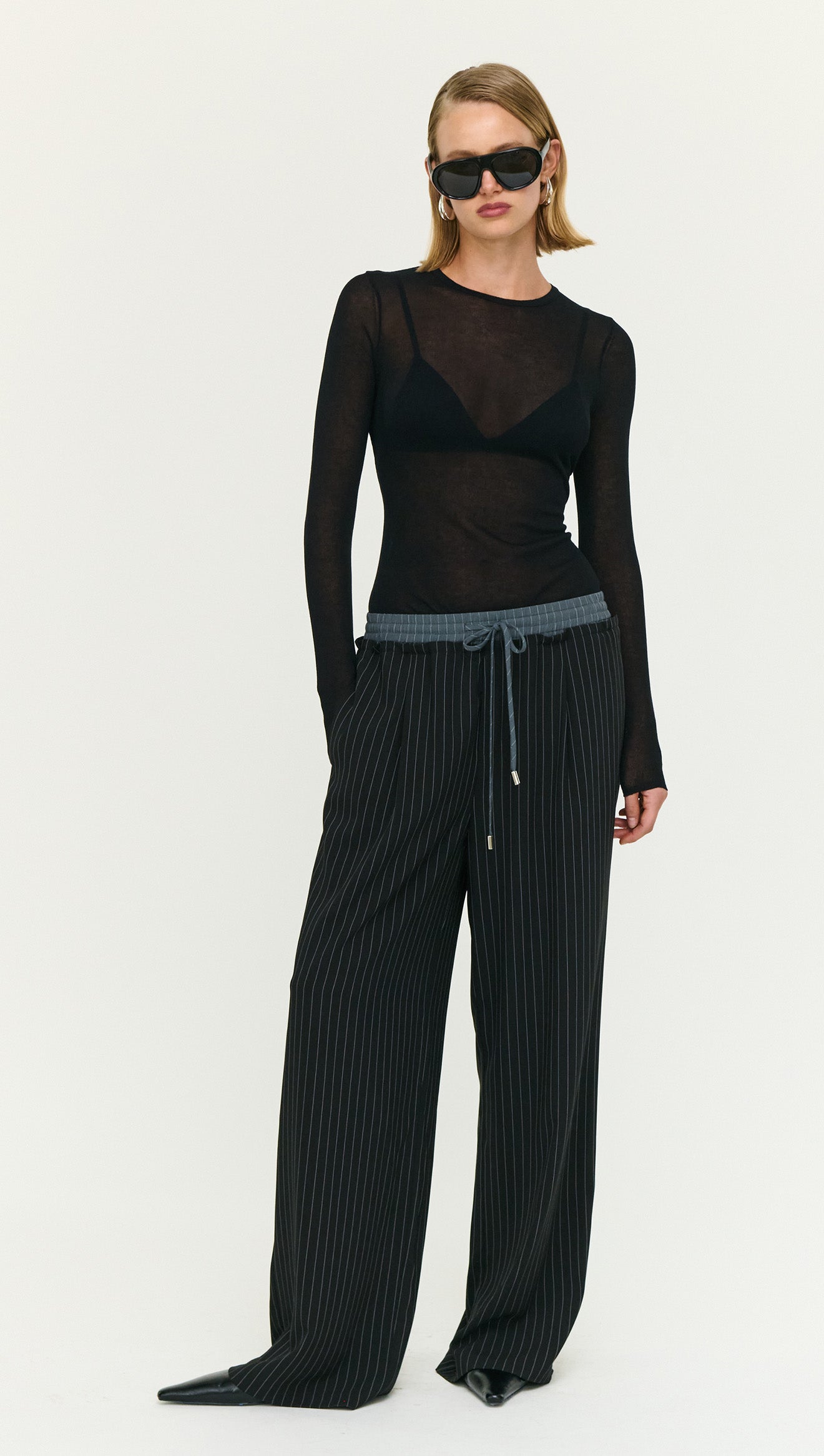 Model wearing Pinstripe Relaxed Drawstring Pants paired with a sheer black top and sunglasses, holding a small black bag.