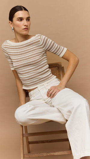 Woman wearing a beige and white striped boat neck knit top, styled casually with wide-leg white pants and sitting on a chair.