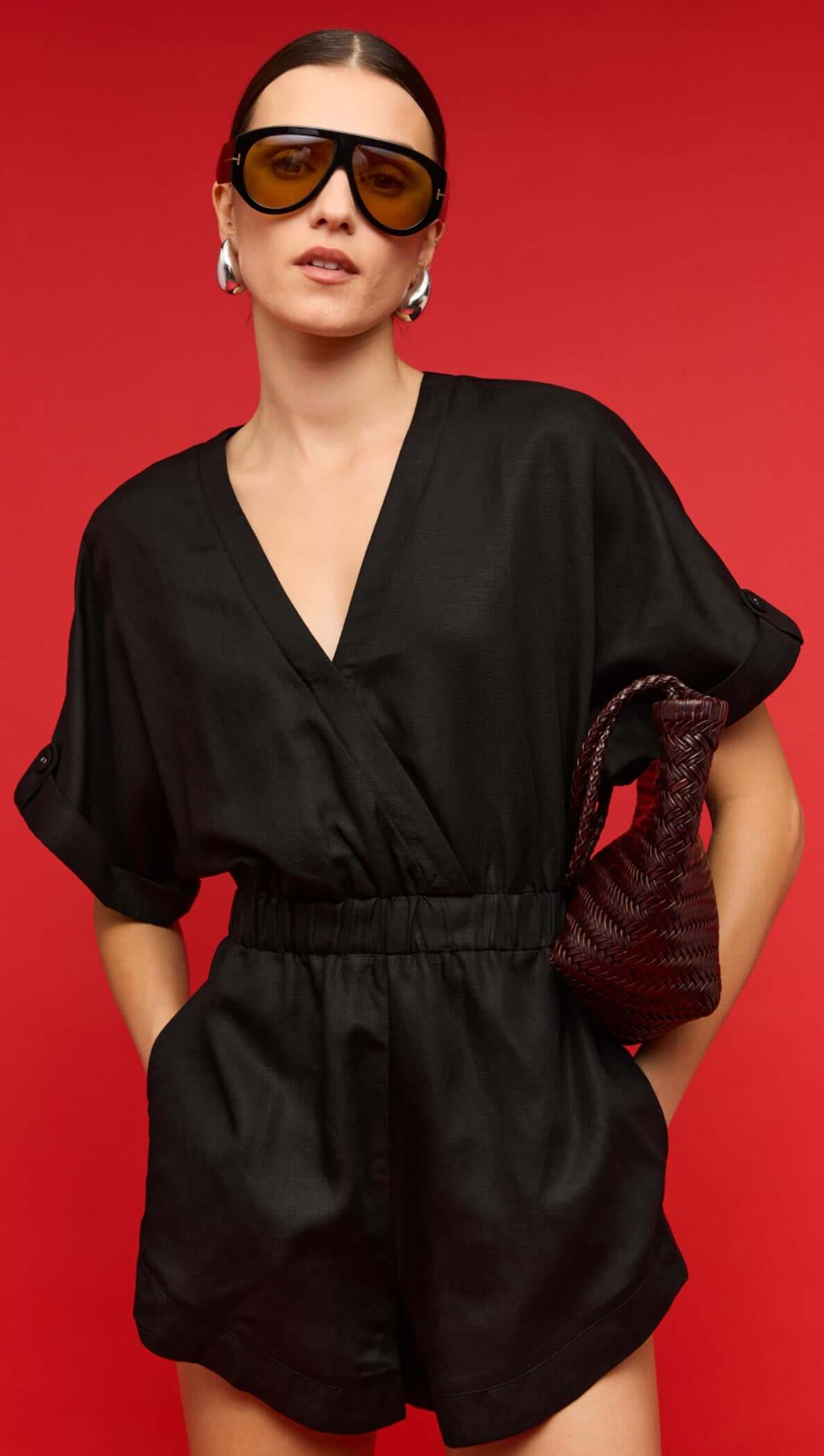 Model wearing a black linen blend V-neck playsuit with sunglasses and a handbag against a red background.