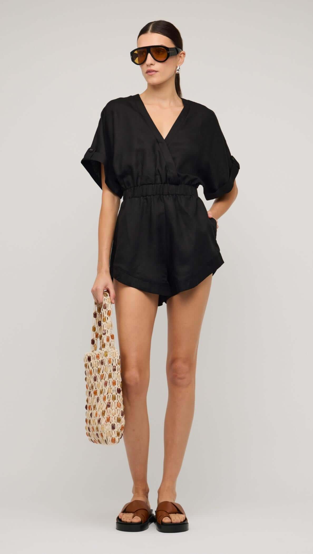 Model wearing a black linen blend V-neck playsuit with an elastic waist, styled with sunglasses and a tote bag.