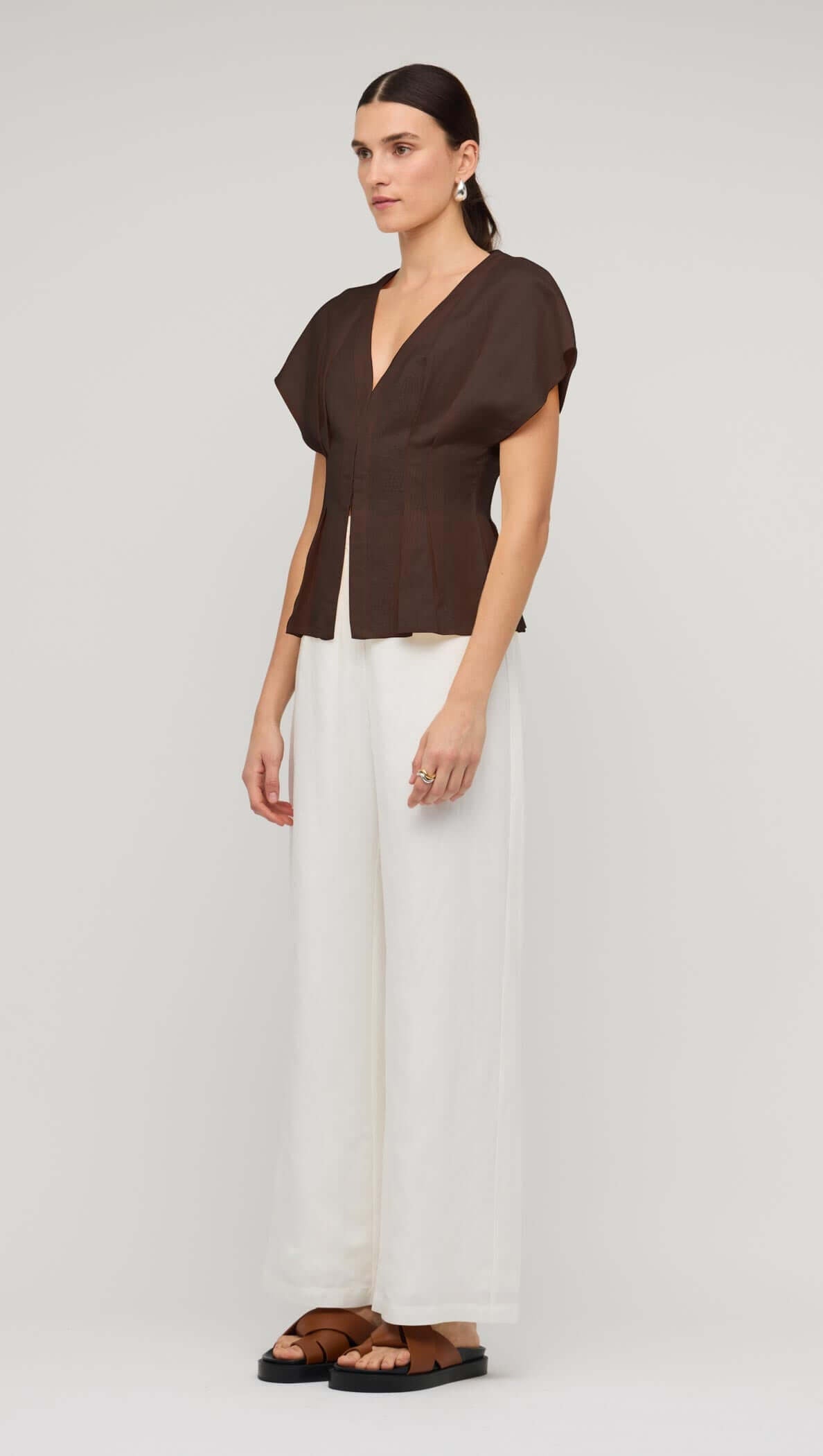 Model wearing a chocolate brown linen pleated V-neck blouse with white wide-leg pants, showcasing a stylish and elegant outfit.