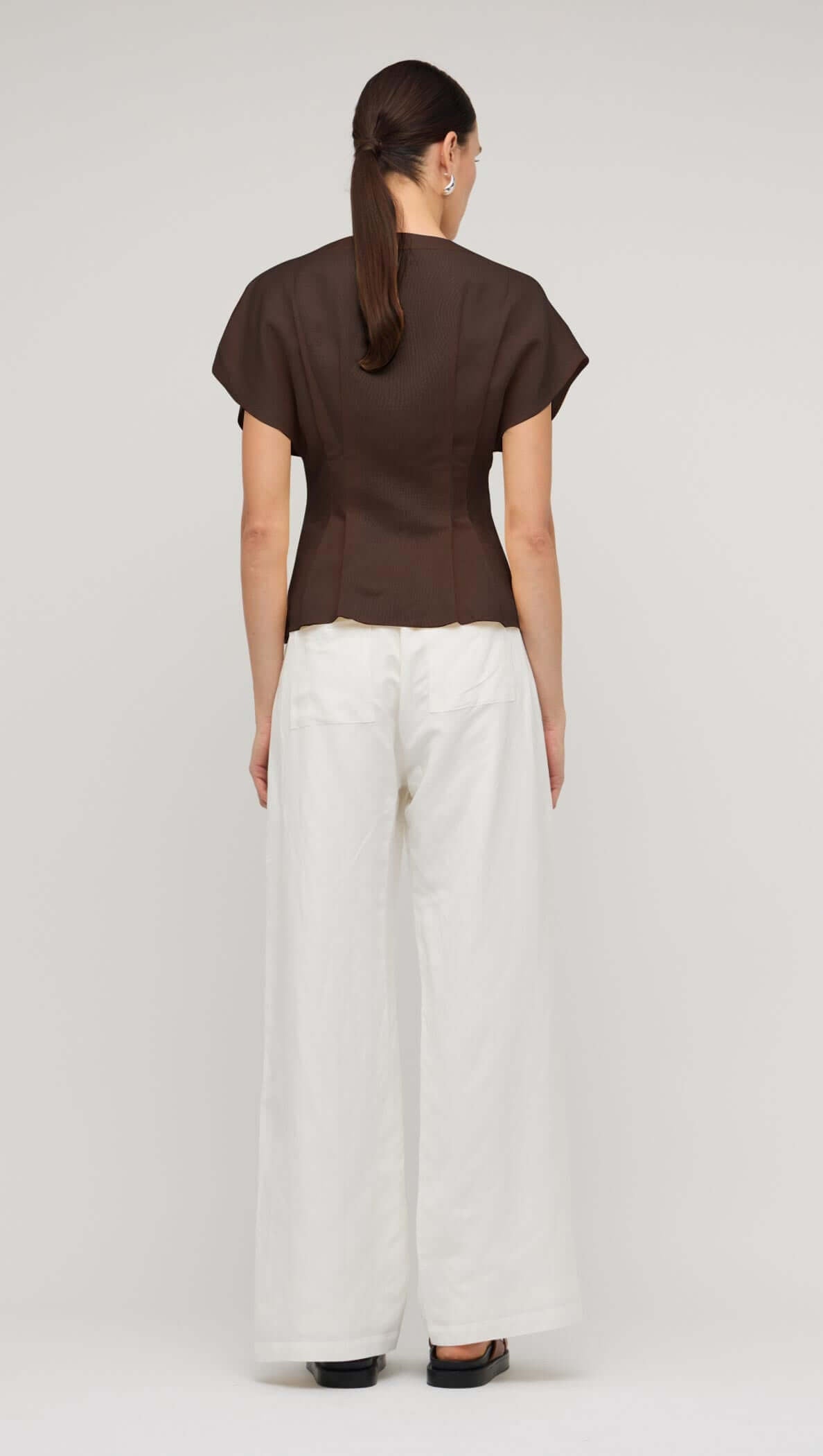 Back view of a chocolate brown linen pleated V-neck blouse paired with white wide-leg pants.