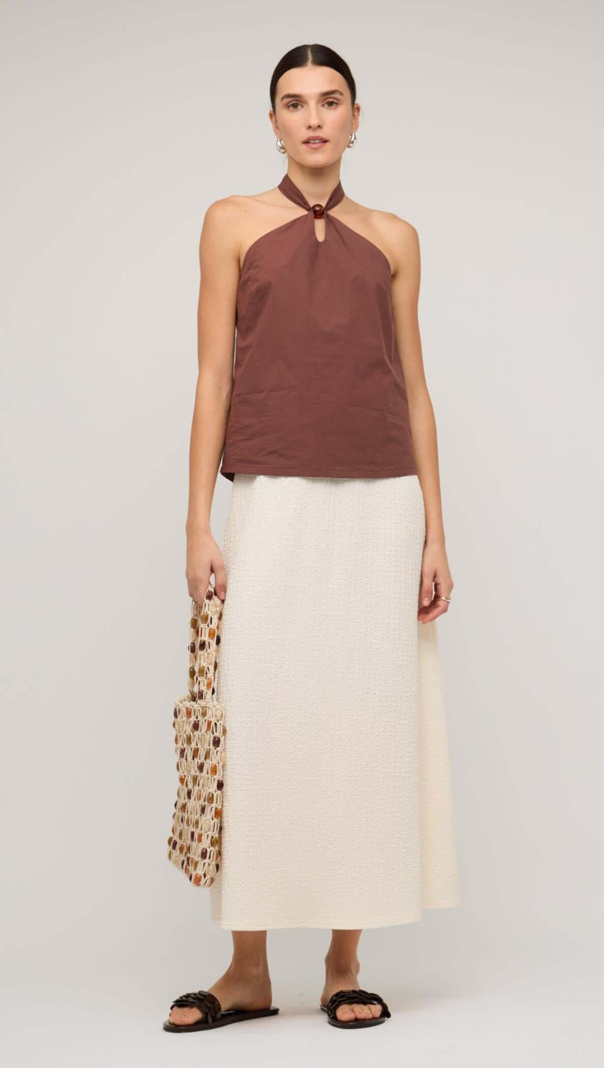 Woman wearing a brown halter top and cream A-line midi skirt, holding a patterned tote bag.