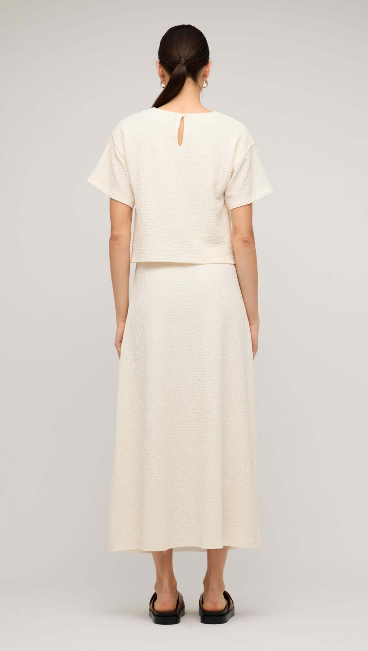 Back view of a model wearing a cream boxy short sleeve cropped top paired with a matching long skirt.