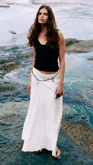 Model wearing a Reversible Black Knit Tank Top with a white skirt near a rocky shoreline.
