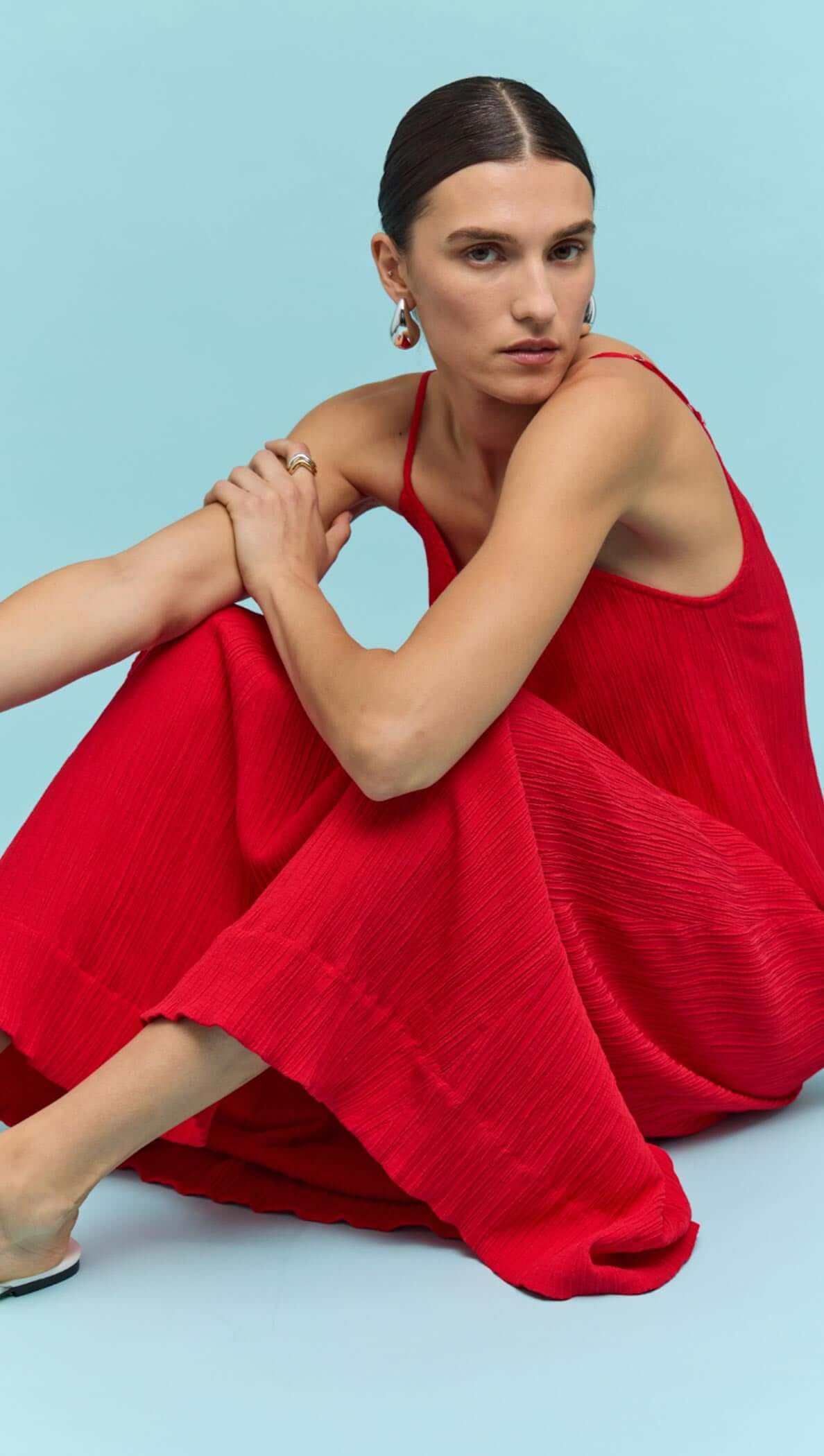 Model sits elegantly in a red crepe V-neck maxi dress against a blue background, showcasing its flowy design.