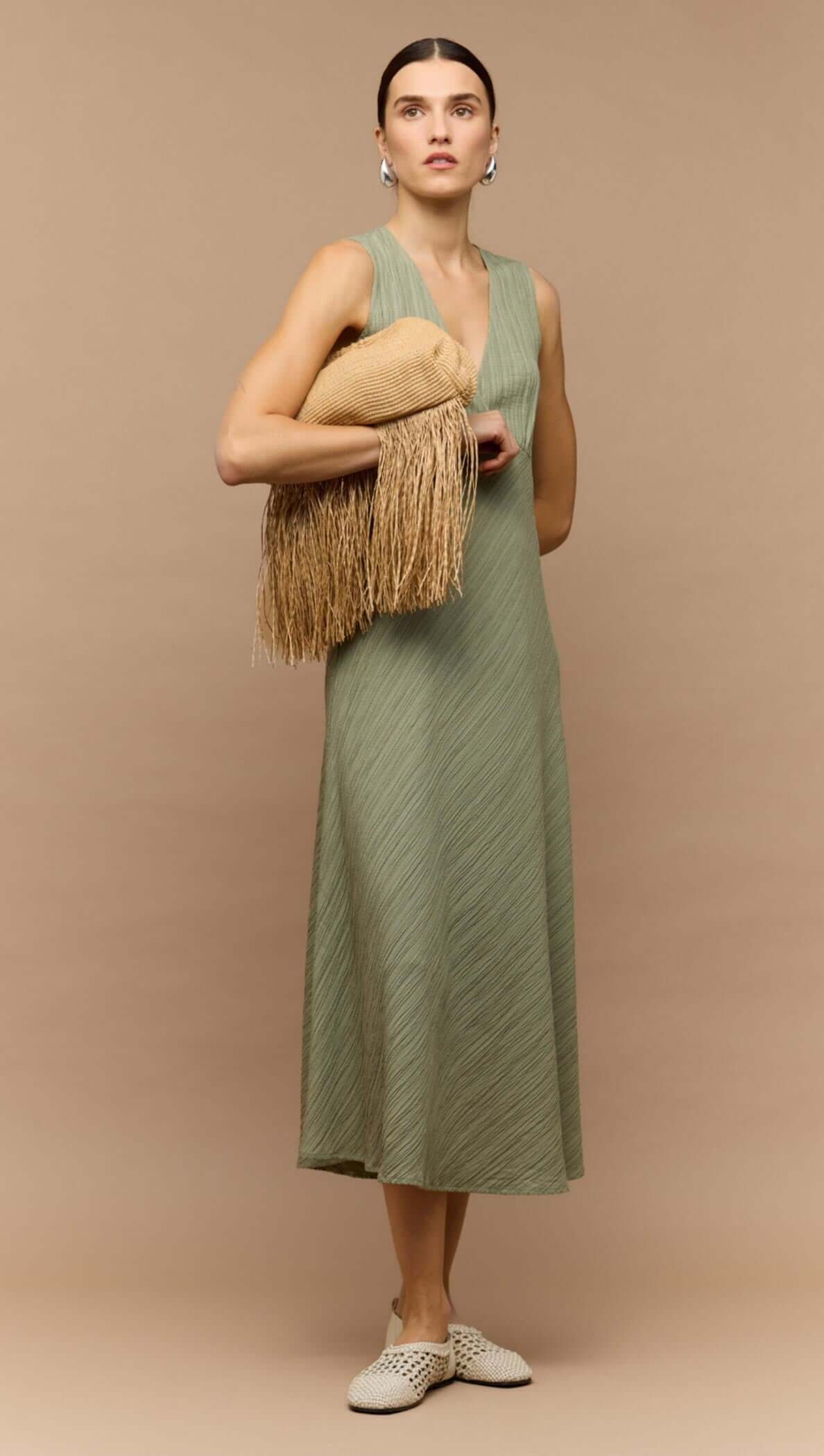 Model wearing a sage green v-neck bias midi dress, holding a fringed handbag, showcasing an elegant style.