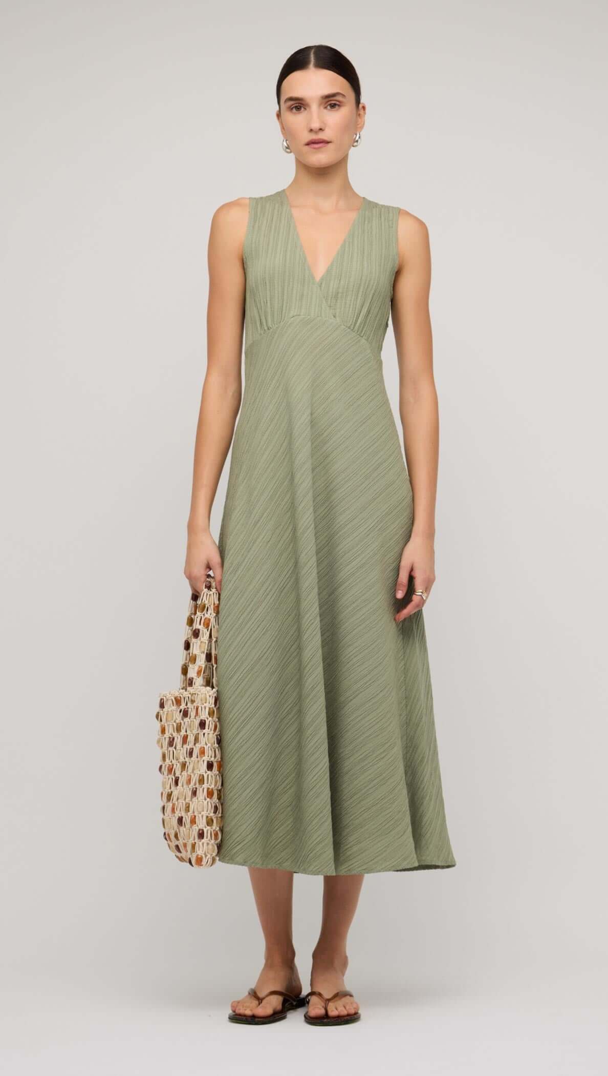 Sage green V-neck bias midi dress worn by model, styled with a patterned tote bag and sandals.