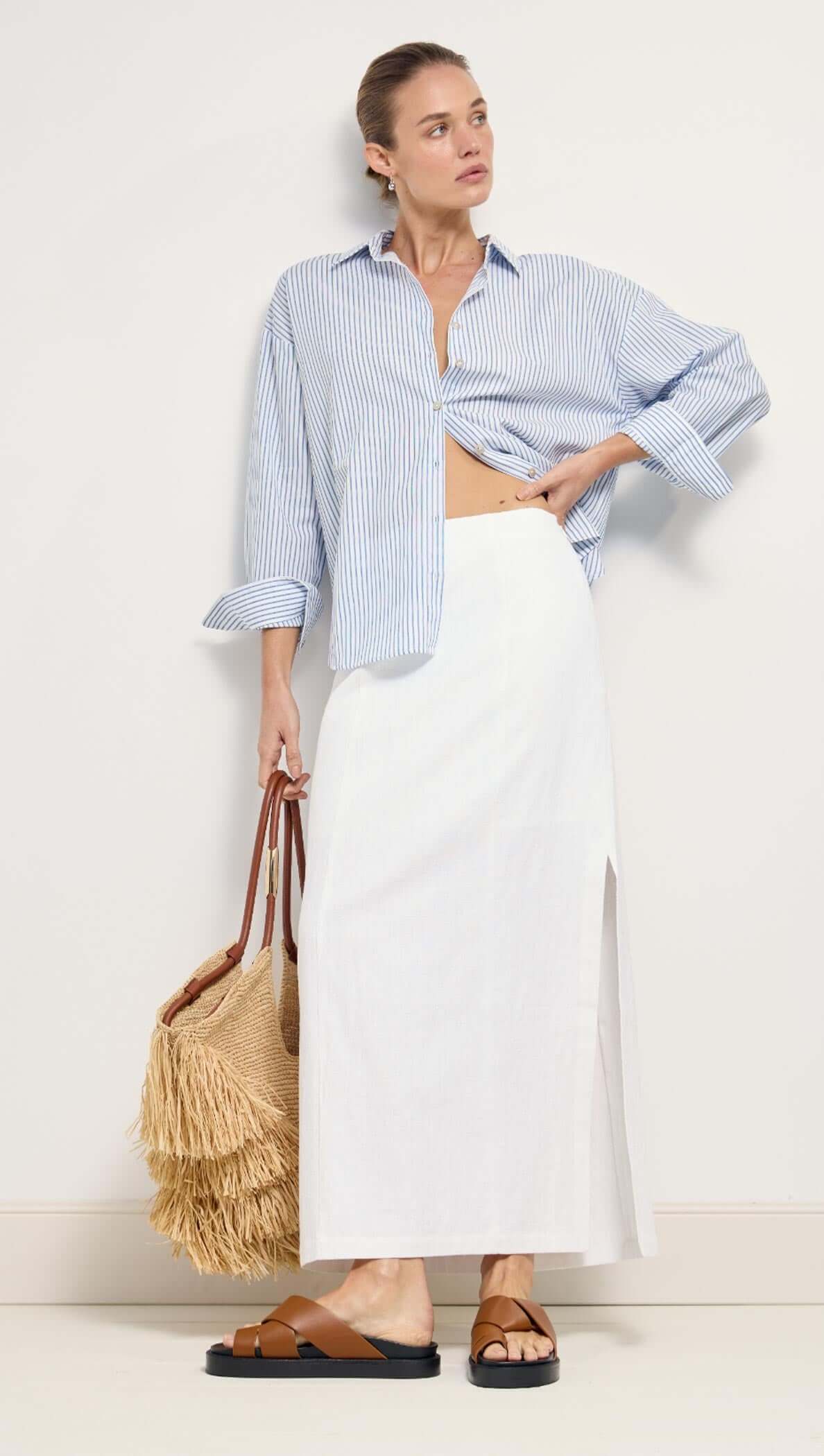 Model wearing a Cotton Blue & White Oversized Shirt with a white skirt and straw bag, showcasing resort casual style.