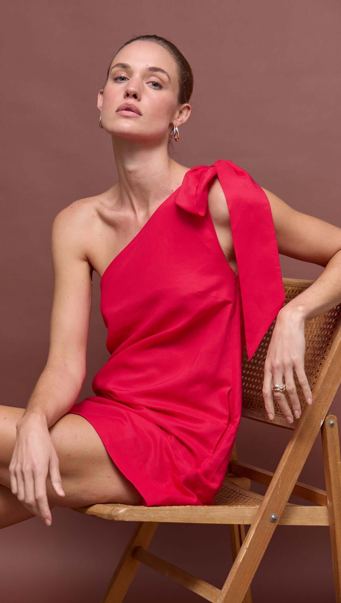 Red Linen Blend One Shoulder Mini Dress worn by a model seated on a chair with a modern and fashionable look.