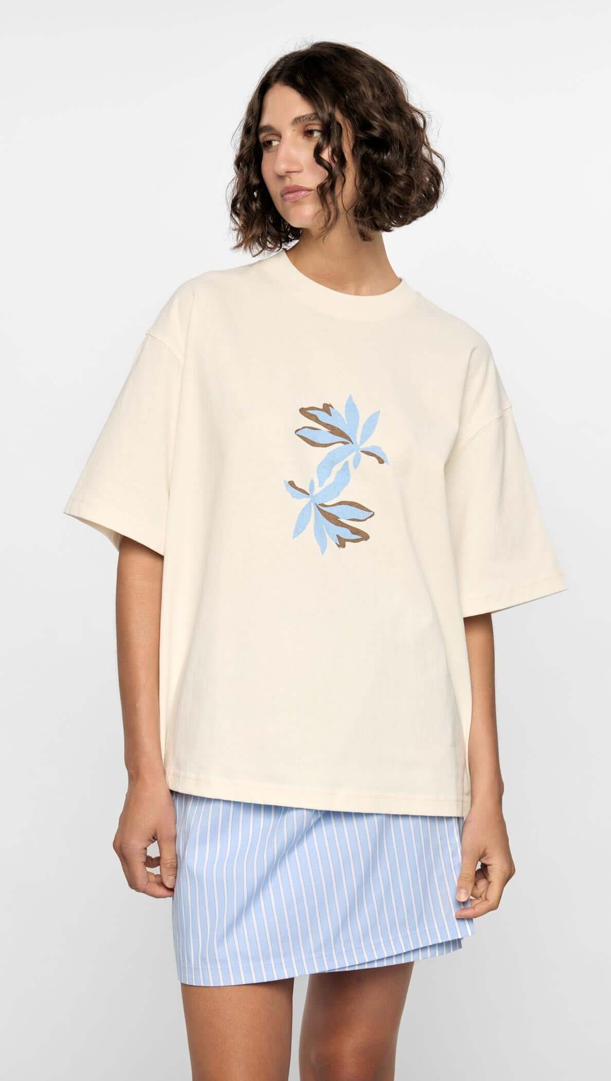 Model wearing Mae Graphic Tee with cream base and blue floral print, styled with a striped skirt.
