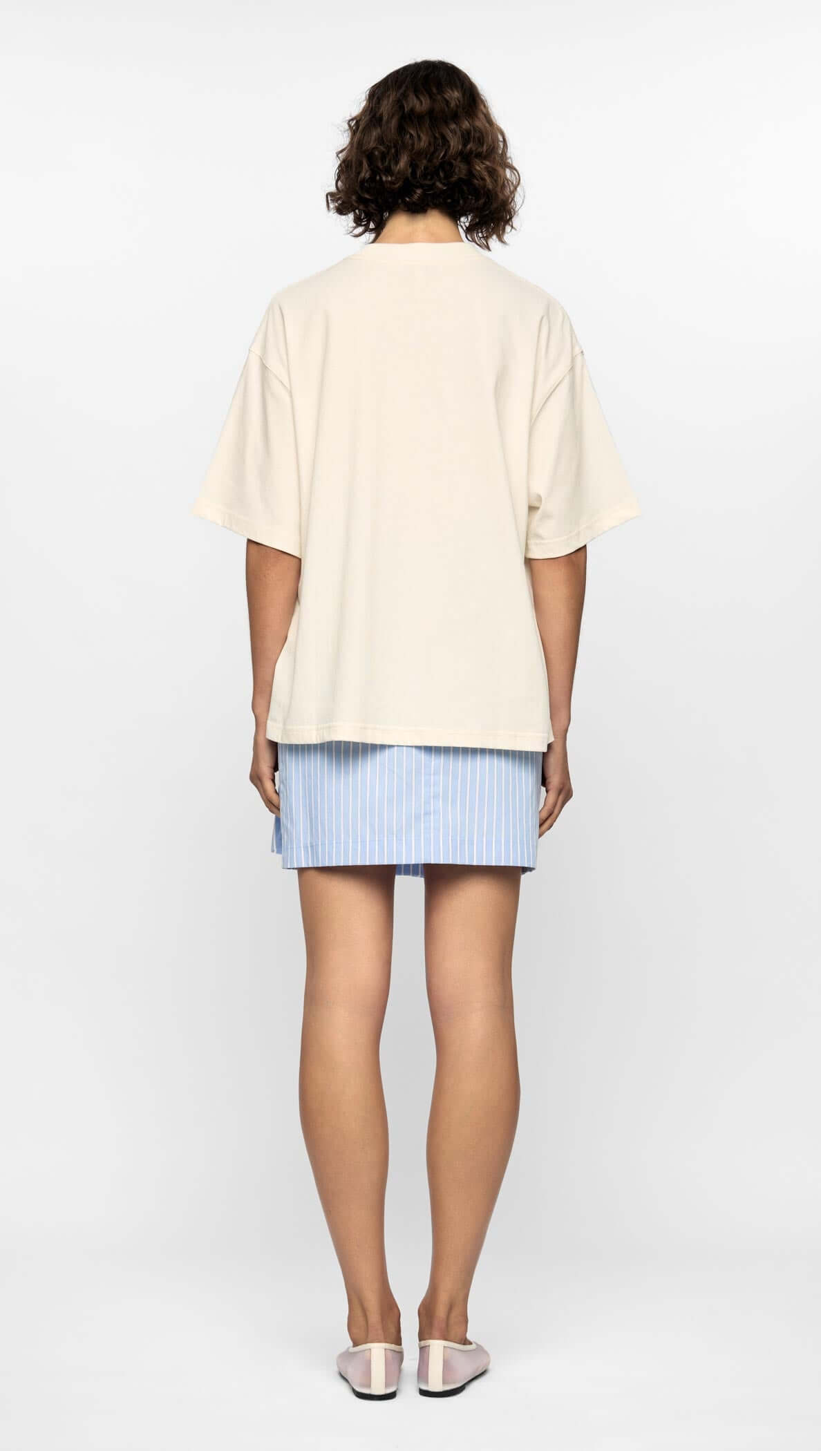 Back view of a model wearing a relaxed cream Mae Graphic Tee with an oversized fit and a blue striped skirt.