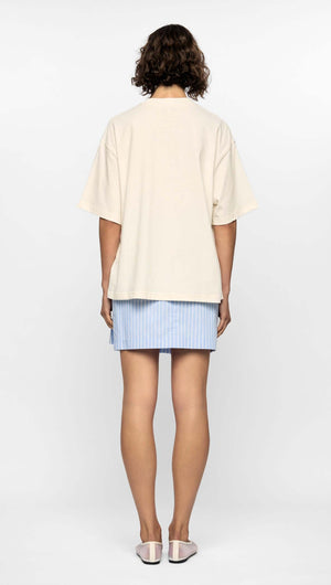 Back view of a model wearing a relaxed cream Mae Graphic Tee with an oversized fit and a blue striped skirt.