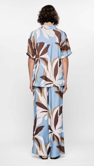 Back view of a woman wearing the Maeve Resort Shirt with bold abstract print in brown, blue, and cream, styled for summer.