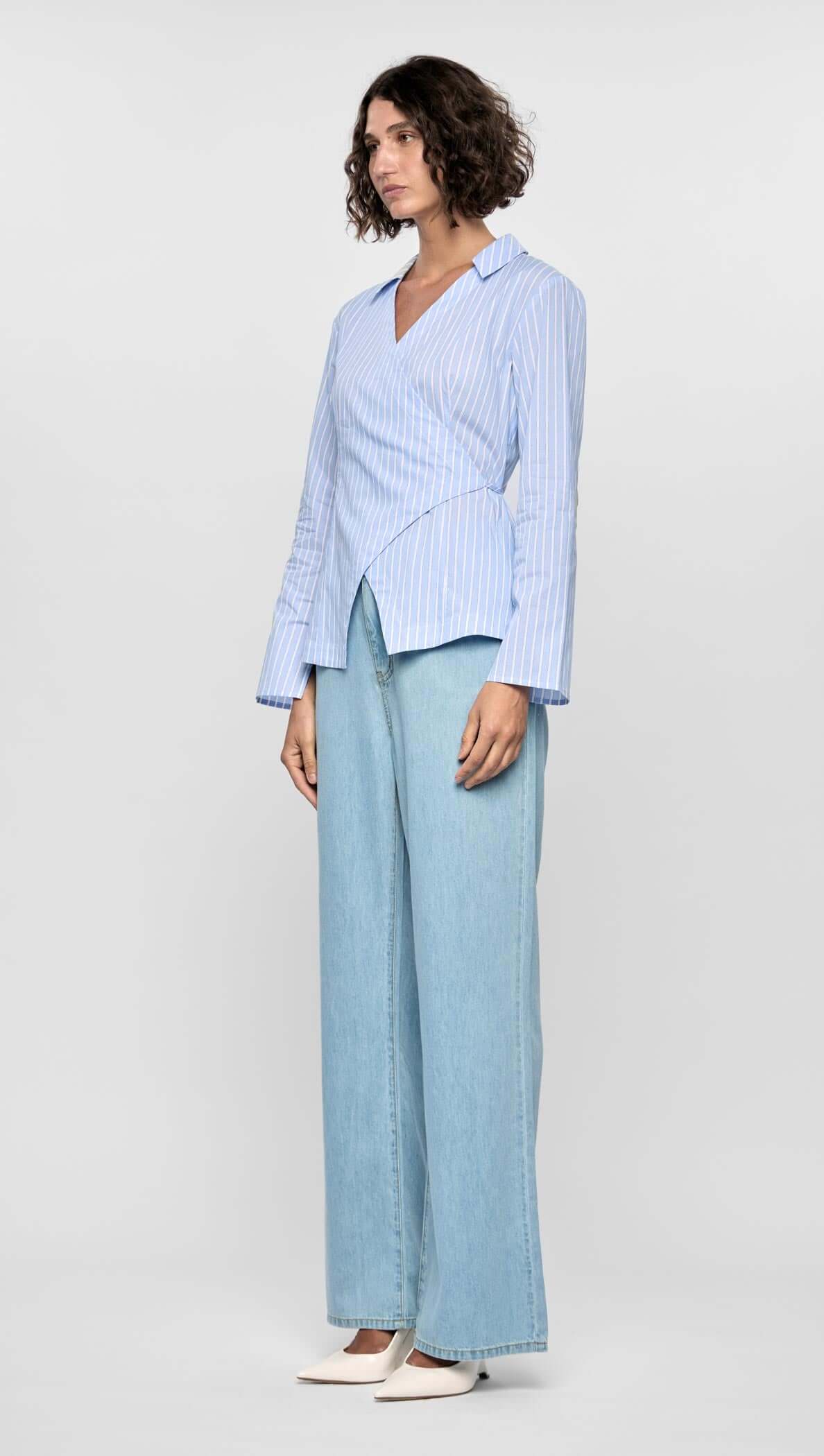 Model wearing the Ayra Pinstripe Wrap Top in light blue, paired with wide-leg jeans, showcasing an asymmetric wrap design.