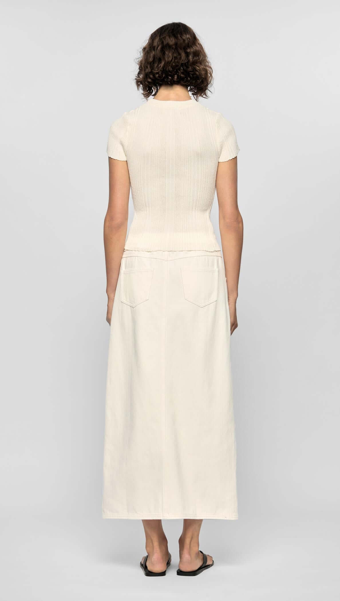 Back view of a model wearing the off-white Emmie Denim Midi Skirt with a fitted top, showcasing its high-waisted design and back pockets.