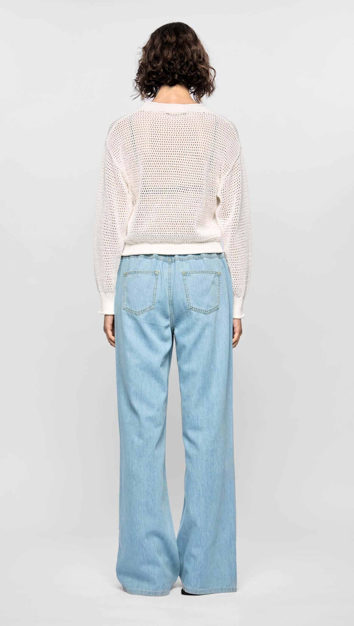 Back view of a model wearing an off-white open-knit Oslo Knit Jumper with relaxed fit, paired with light blue wide-leg jeans.