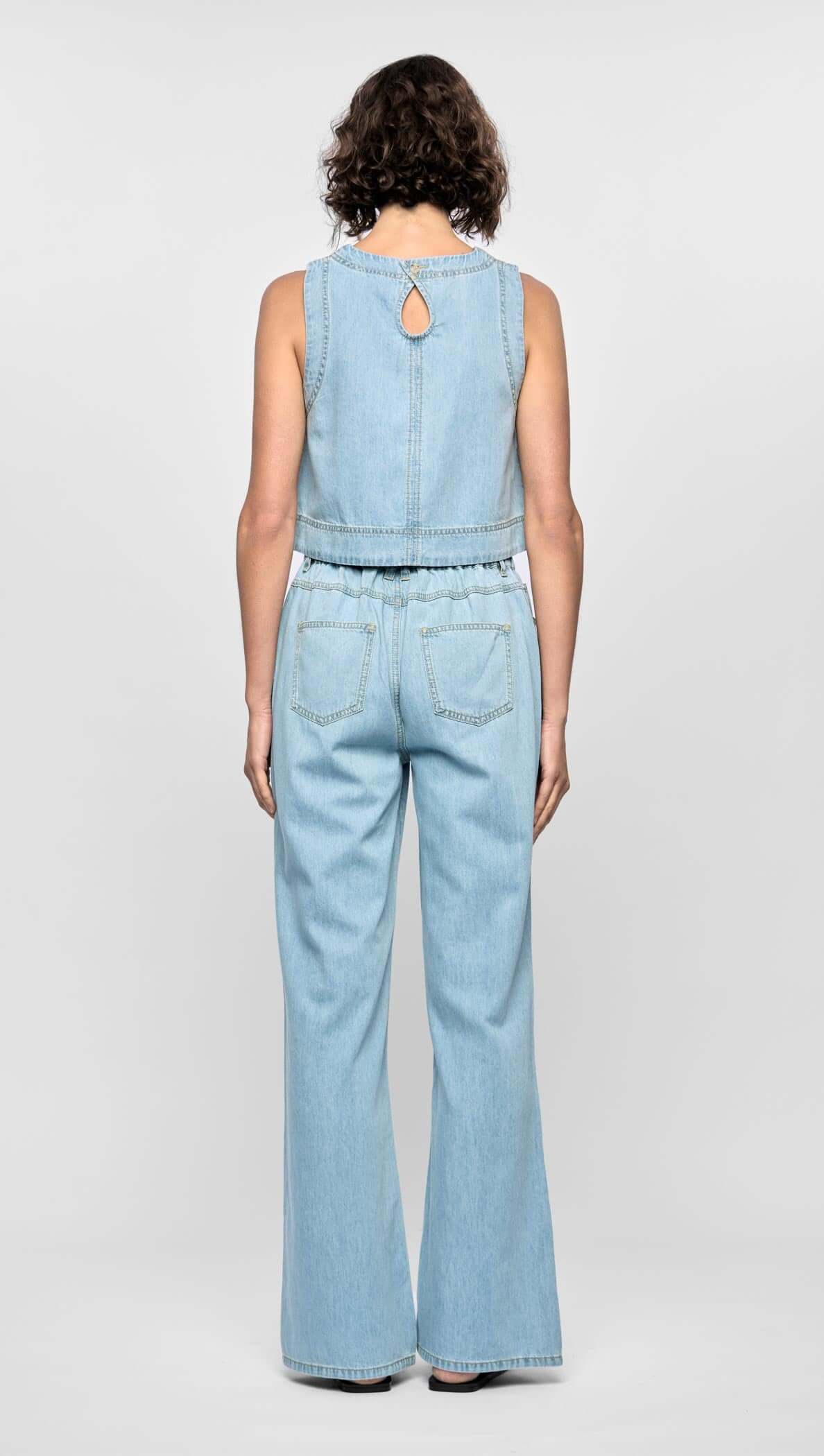 Back view of a woman wearing a light chambray blue sleeveless top with a keyhole button closure and matching jeans.