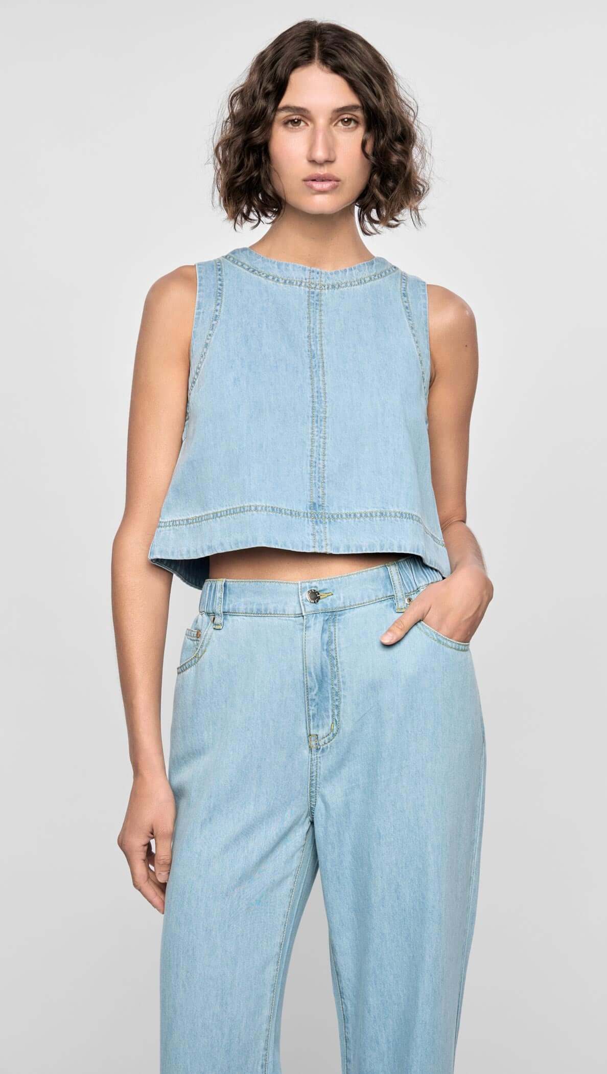 Model wearing the Amelia Chambray Top, a cropped, sleeveless top in light chambray blue with raw-edge detailing.