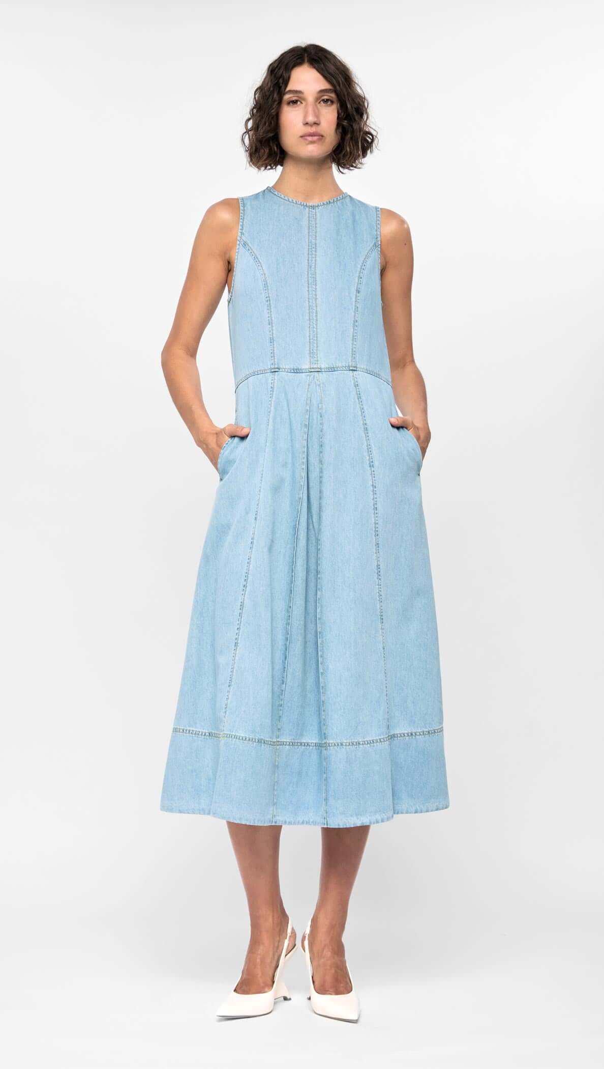 Model wearing the Amelia Chambray Midi Dress, a light blue sleeveless dress with a fitted bodice and side pockets.