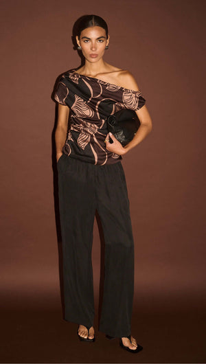 Abstract Leaf Print One Shoulder Top modeled with black wide-leg pants and a clutch on a brown background.