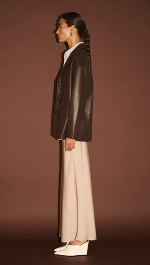 Model wearing a Chocolate Relaxed Vegan Leather Blazer with a white shirt and light pants against a brown background.