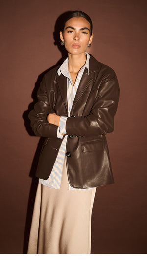 Model wearing a Chocolate Relaxed Vegan Leather Blazer over a light shirt, against a brown background.