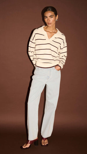 Model wearing a Chocolate Stripe Polo Knit Jumper with light blue jeans against a brown background.