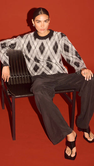 Model wearing a Relaxed Fit Argyle Brushed Jumper seated on a chair against an orange backdrop.
