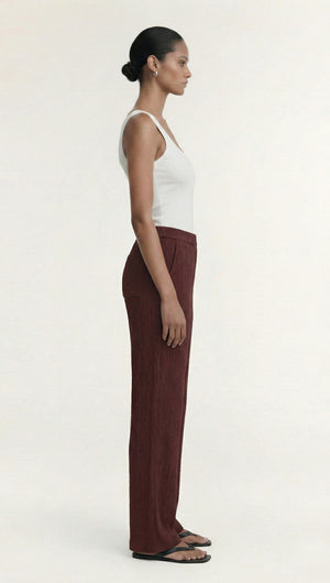 Side view of a model wearing Textured Burgundy Relaxed Fit Pant with a white tank top and sandals.