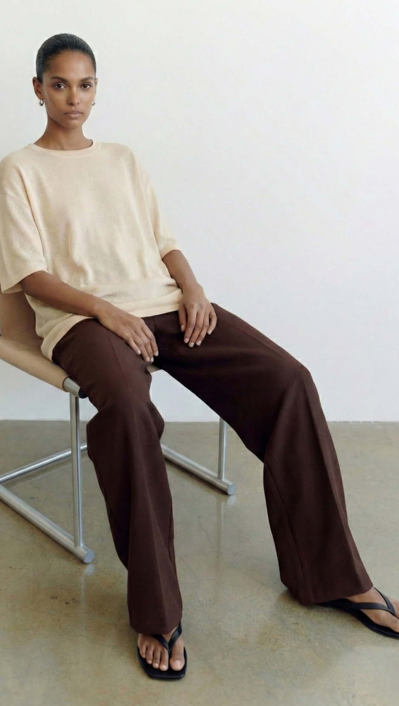 Model wearing a White Round Neck Oversized Knit Tee with brown pants, seated on a chair.