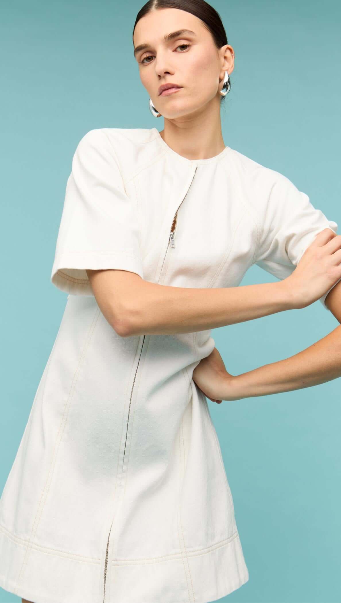 Model wearing a white denim mini dress with front zip and short sleeves against a teal background.