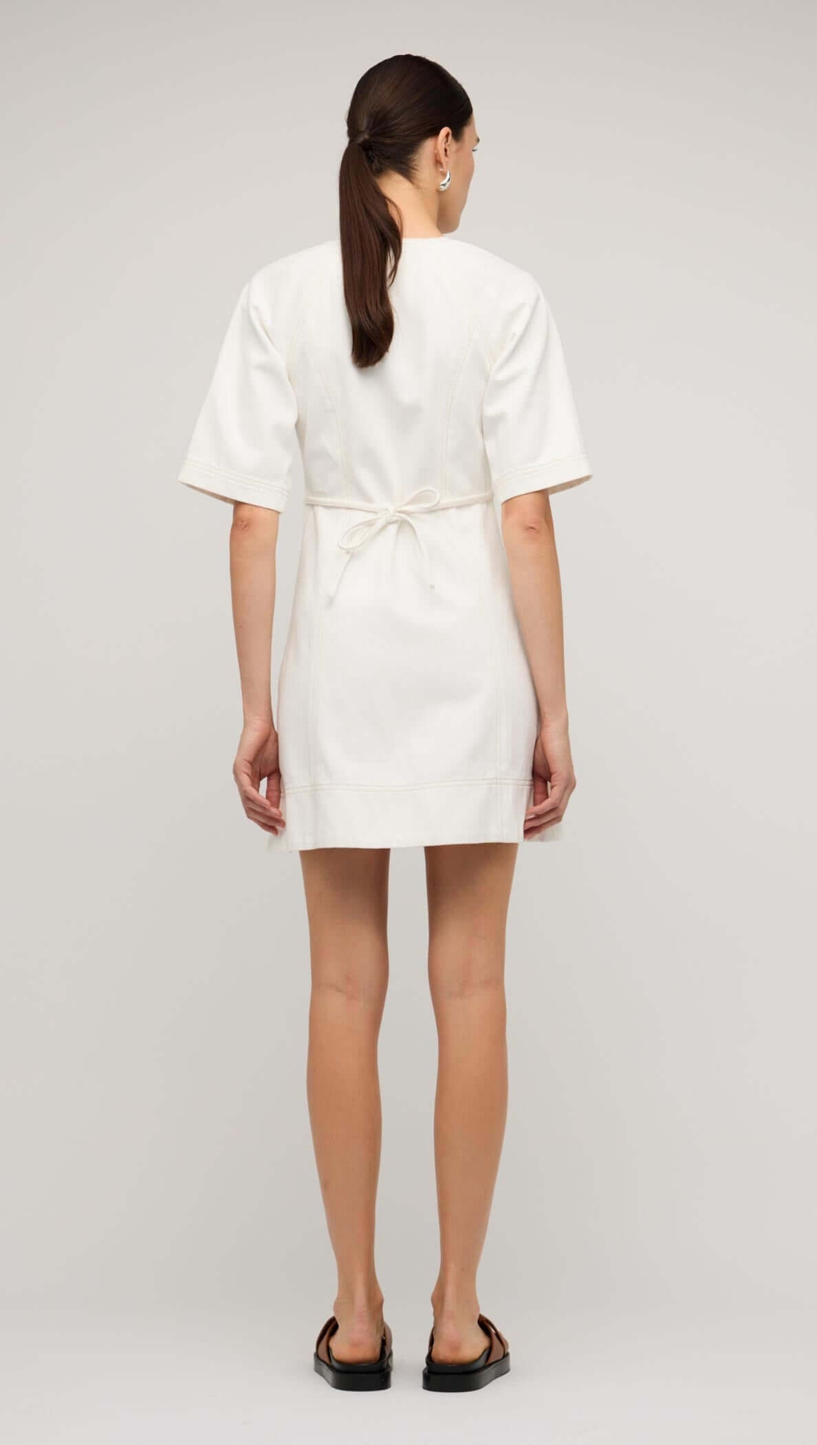 Back view of a woman wearing a white denim mini dress with short sleeves and belt detail.