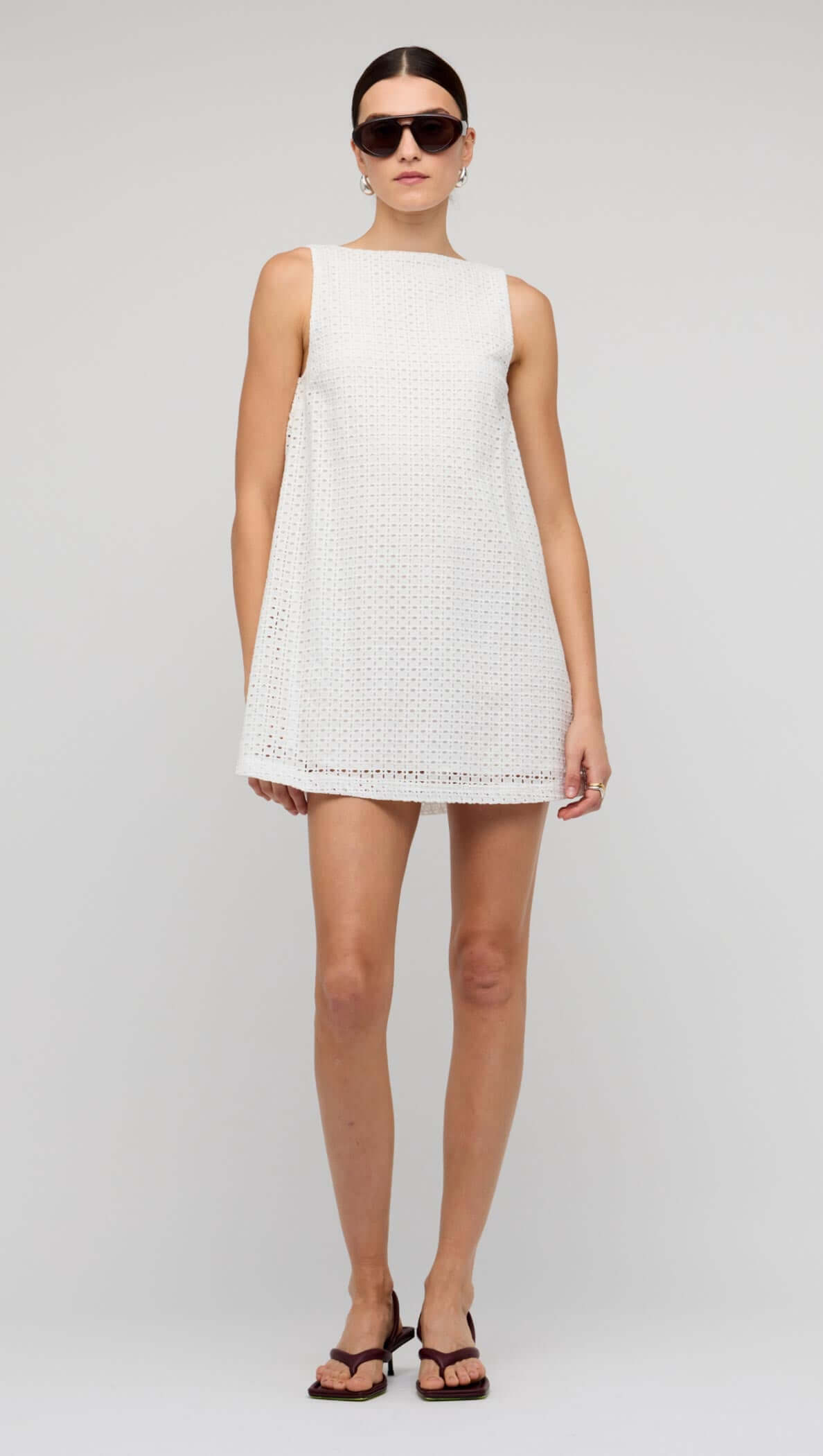 Woman wearing a White Broderie Mini Shift Dress with sunglasses, showcasing a shift silhouette and deep V-back.