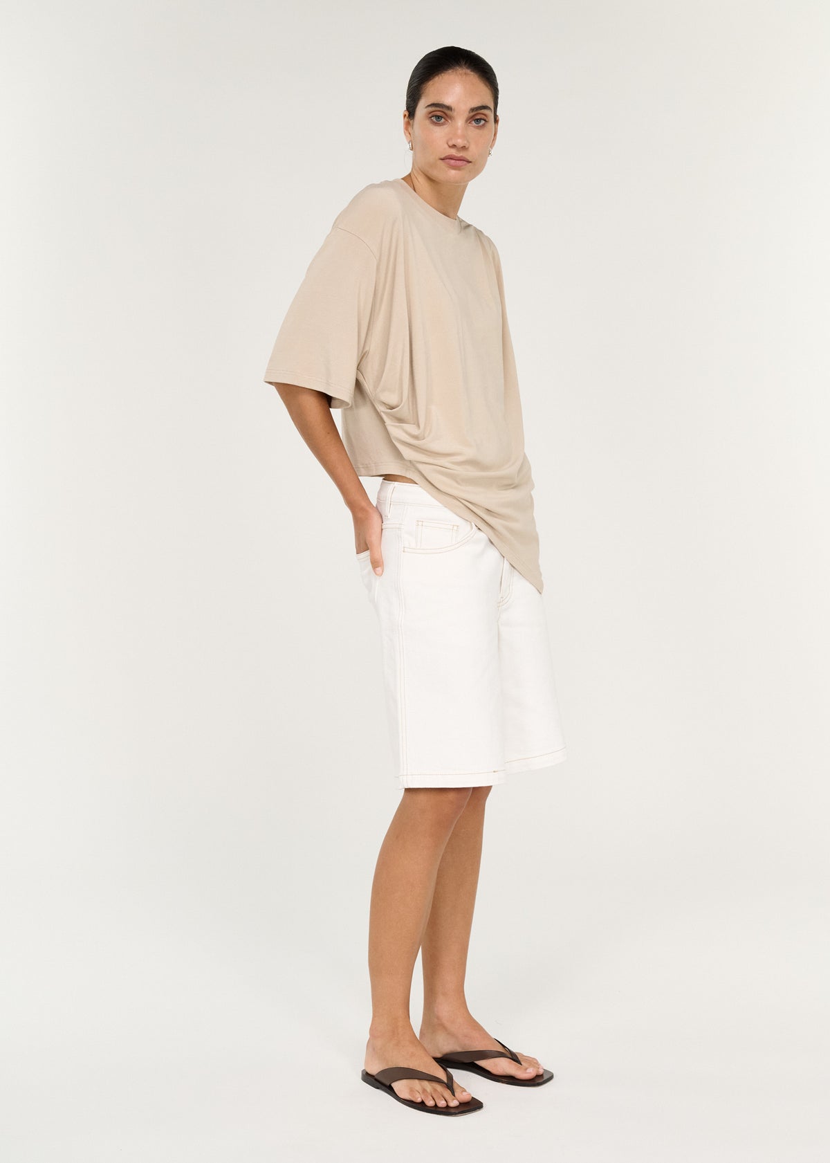 Model wearing Asymmetric Draped Oversize Tee in Beige with dropped shoulders and relaxed short sleeves, turned to the side.