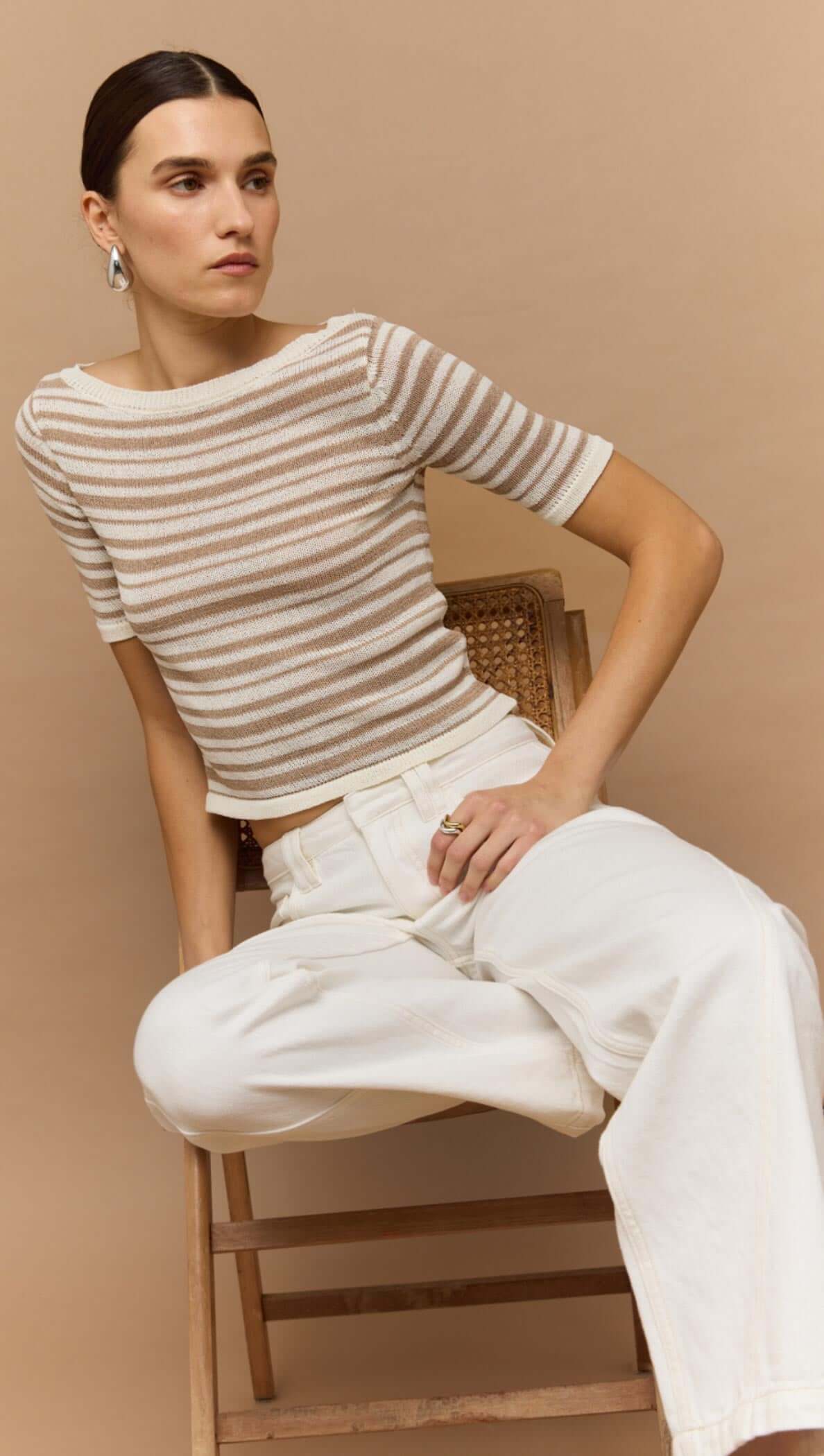 Woman wearing a beige and white striped boat neck knit top, styled casually with wide-leg white pants and sitting on a chair.