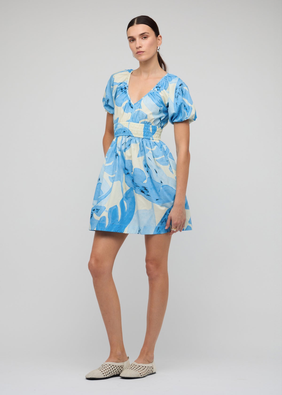 Model wearing Blue and White Floral Cotton V-Neck Mini Dress with a mini length with shirred waistband, turned to the side.