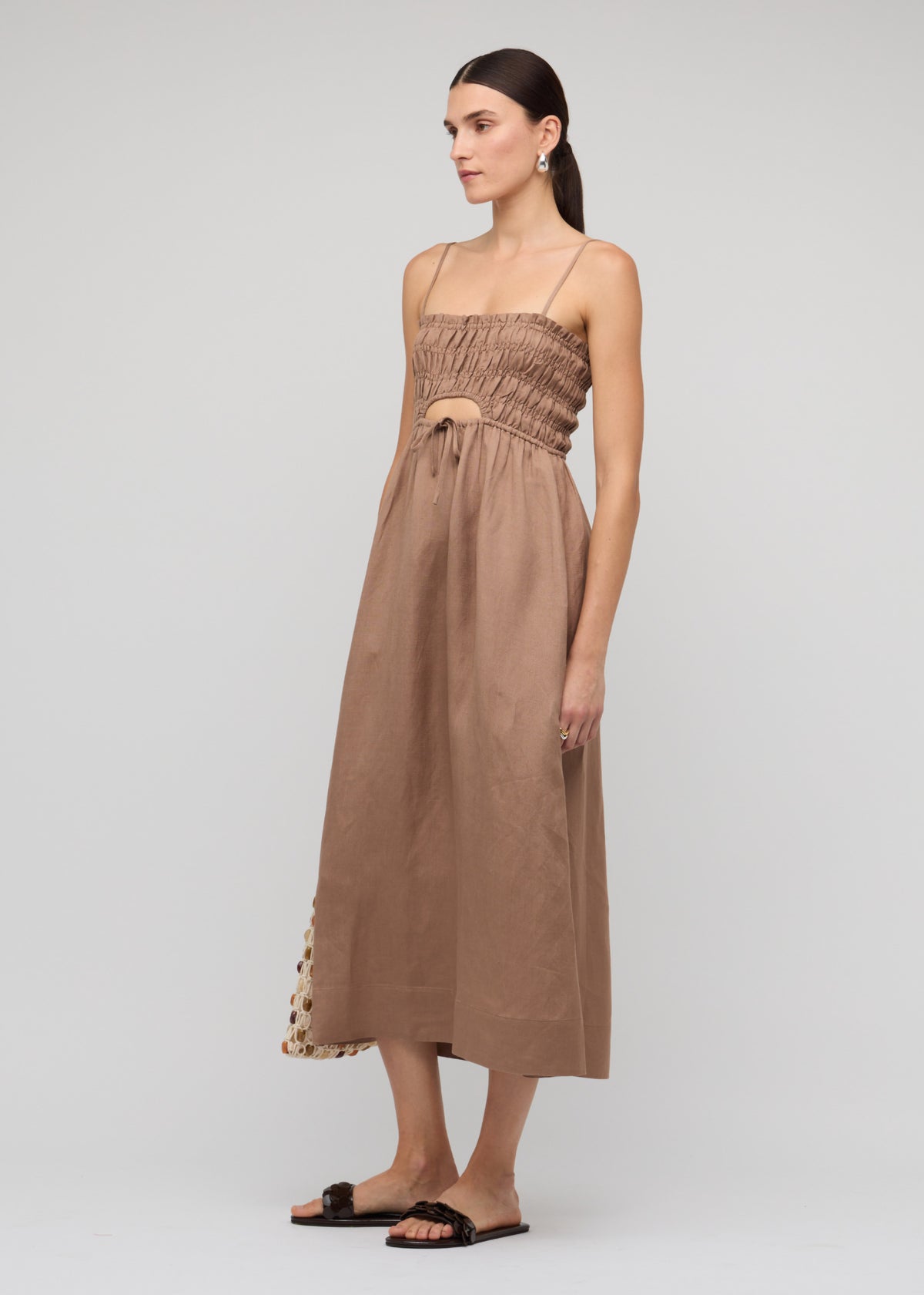 Model wearing Brown Shirred Cut-Out Linen Midi Dress with a shirred fitted bodice with tie-front detail, turned to the side.