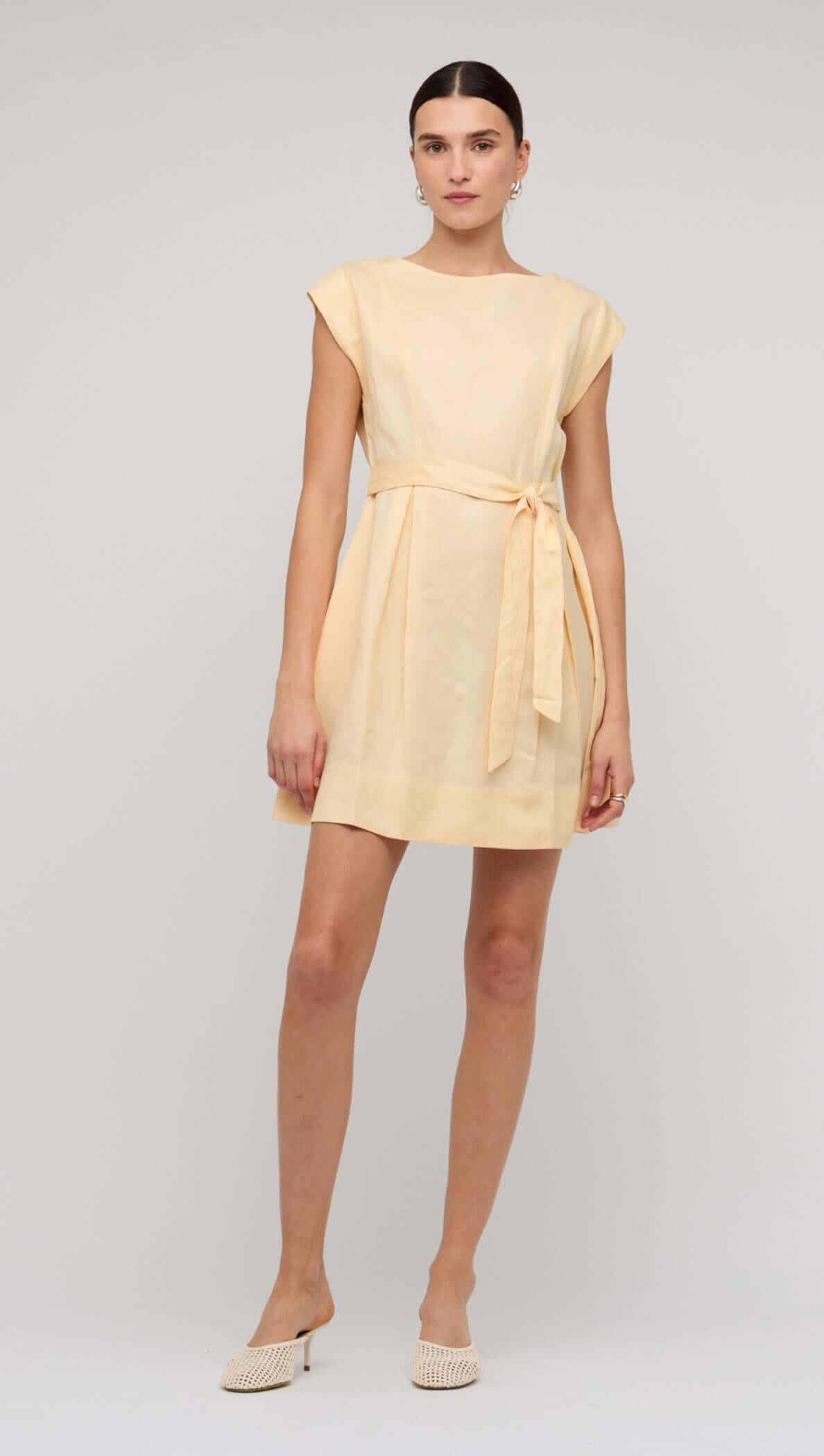 Model wearing a Butter Yellow Linen Mini Dress with cap sleeves and a self-tie belt against a blue background.