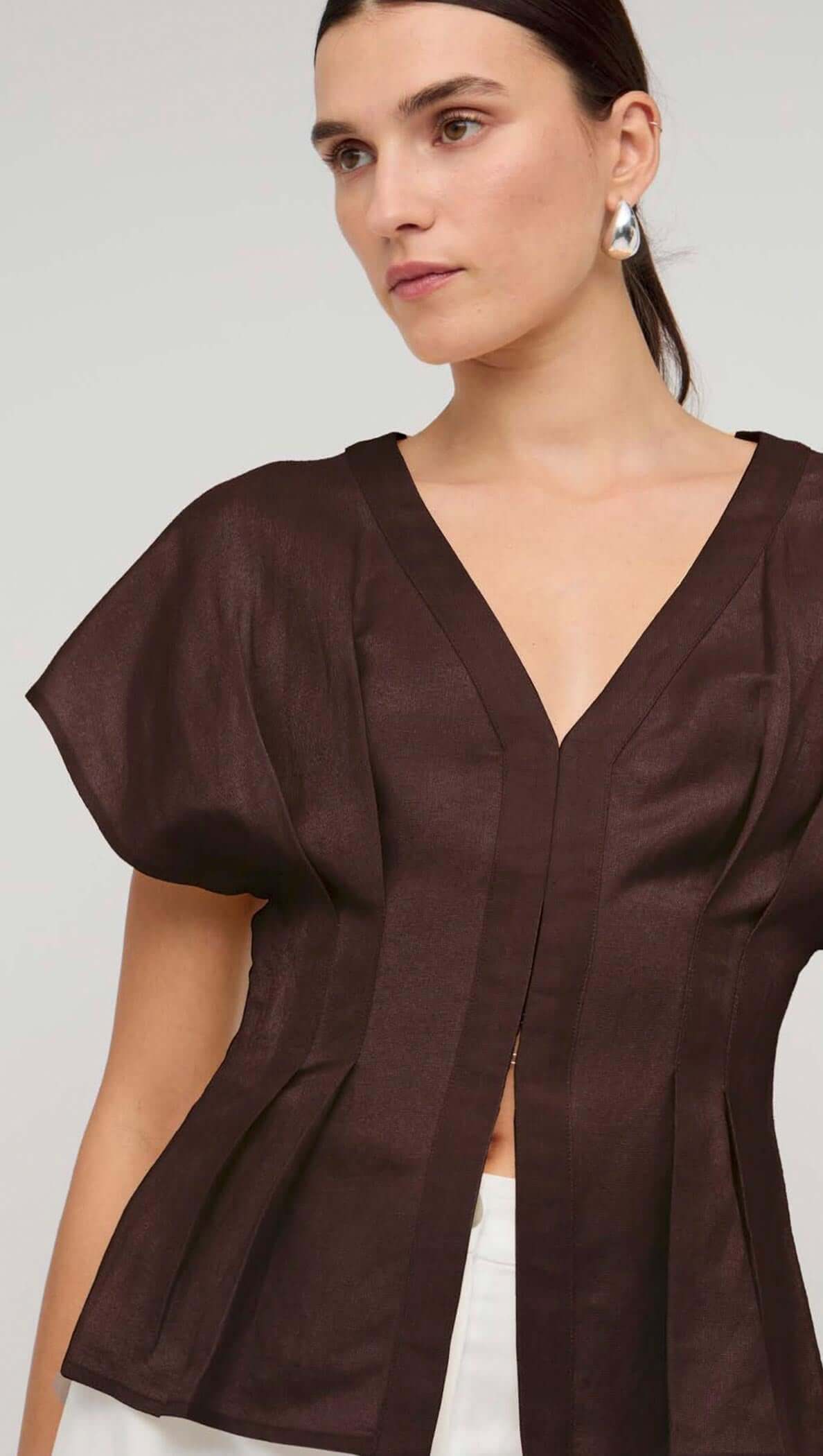 Woman modeling a chocolate brown linen pleated V-neck blouse with structured sleeves.